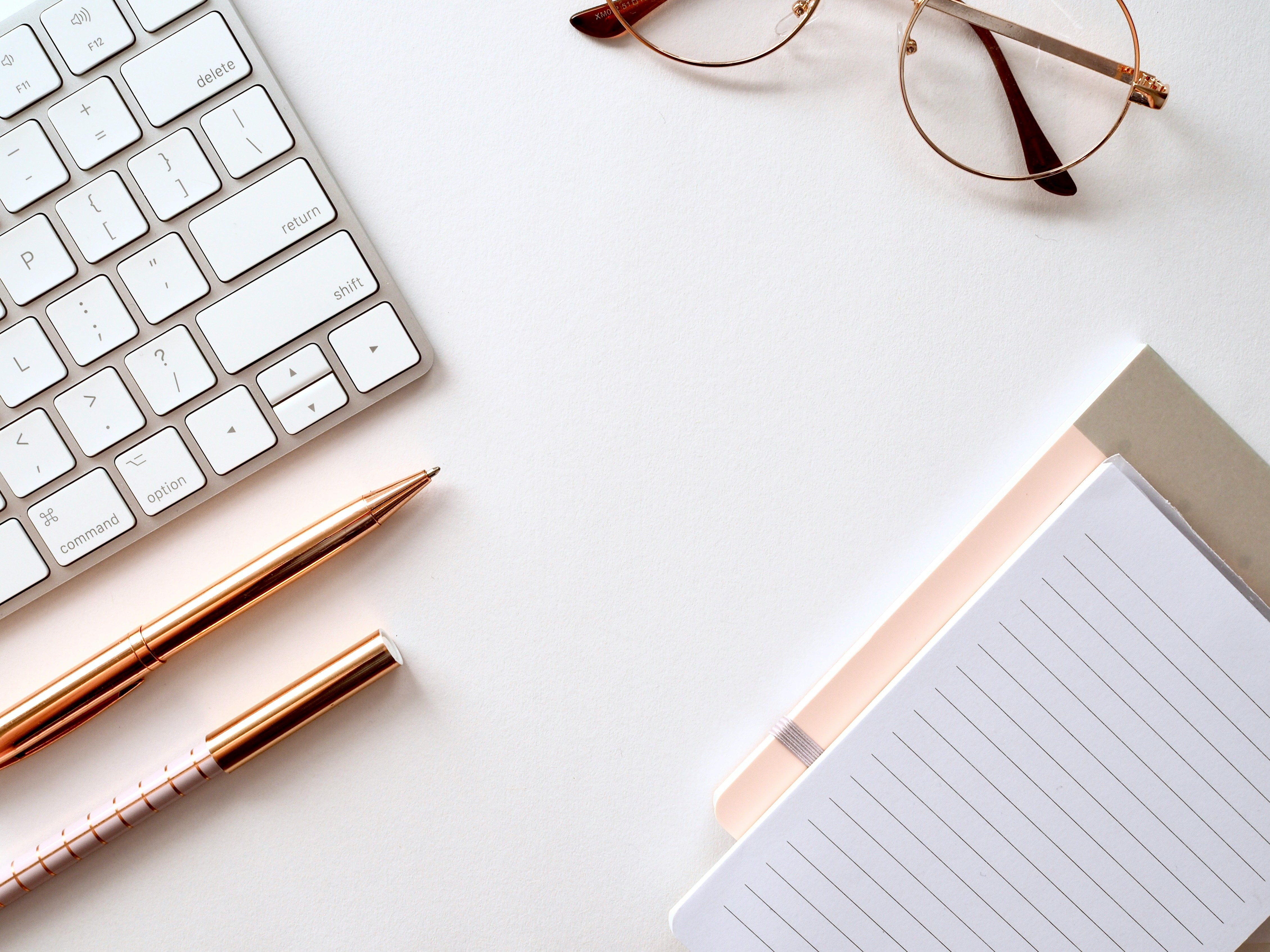 A picture of a keyboard, glasses, pens, and notepad laying on a desk