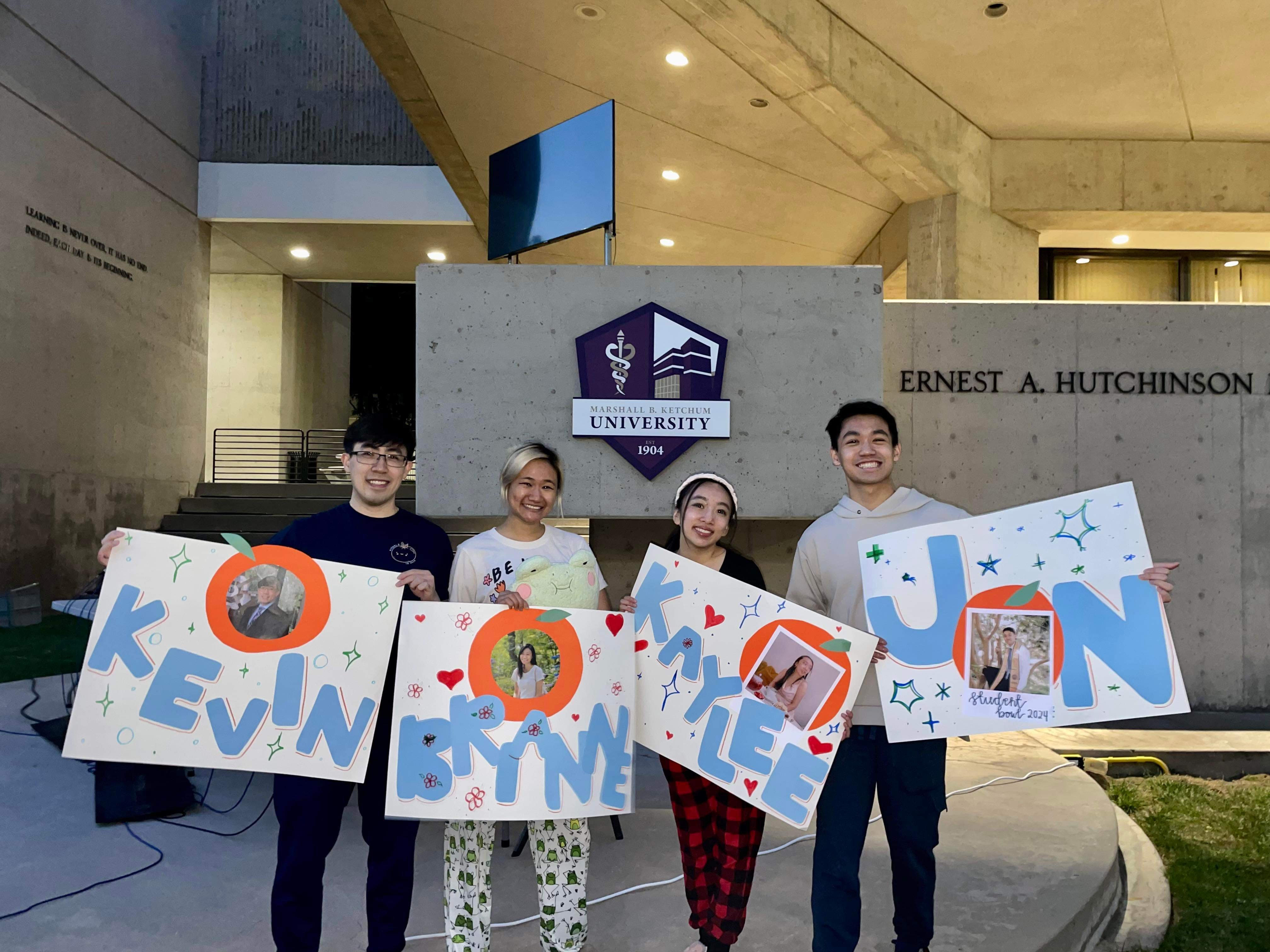 A photo of students standing in front of a building at Marshall B. Ketchum University and holding poster boards with their names and photos on them