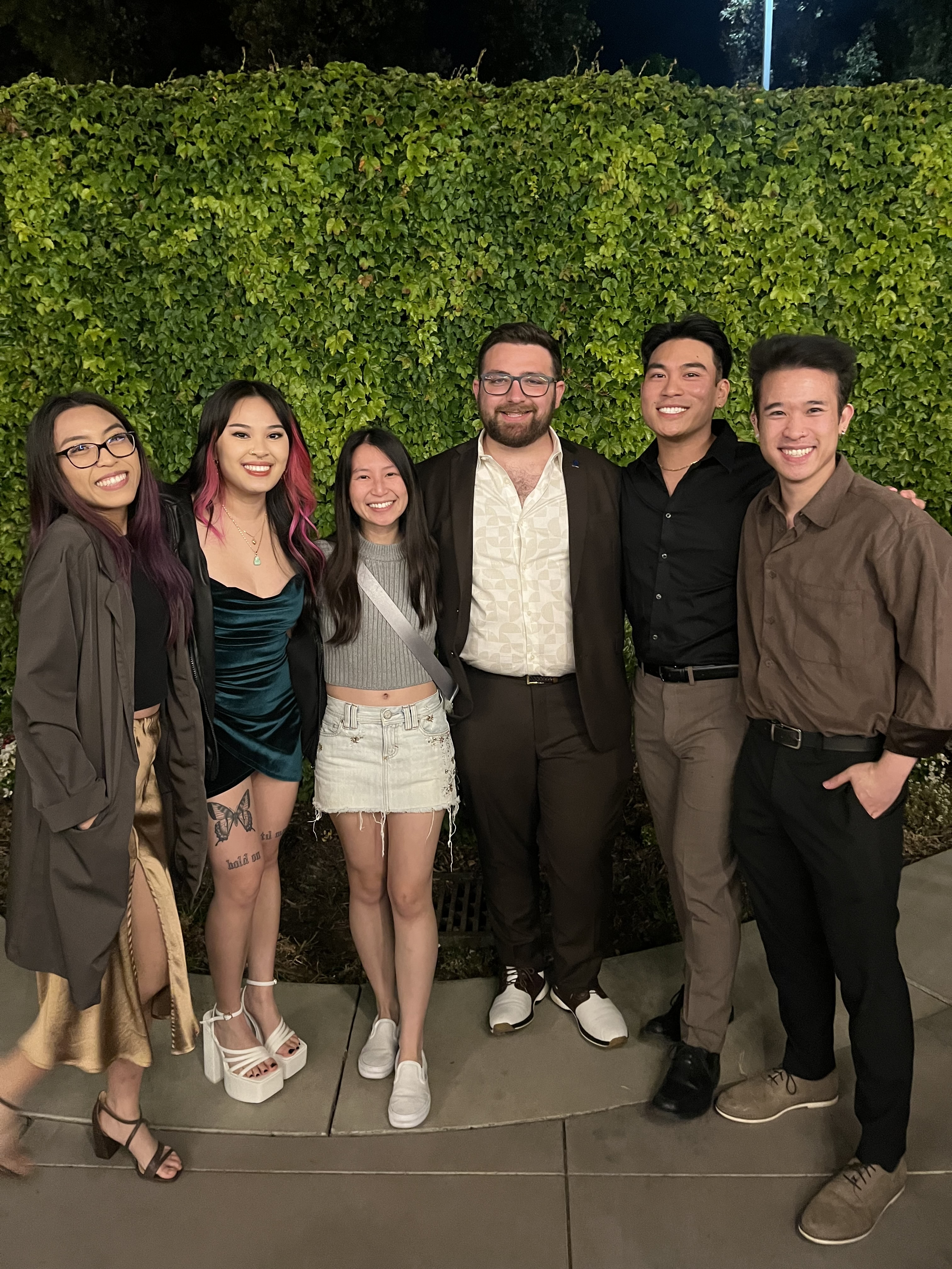 Photo of a group of six people standing together and smiling in front of a hedge.