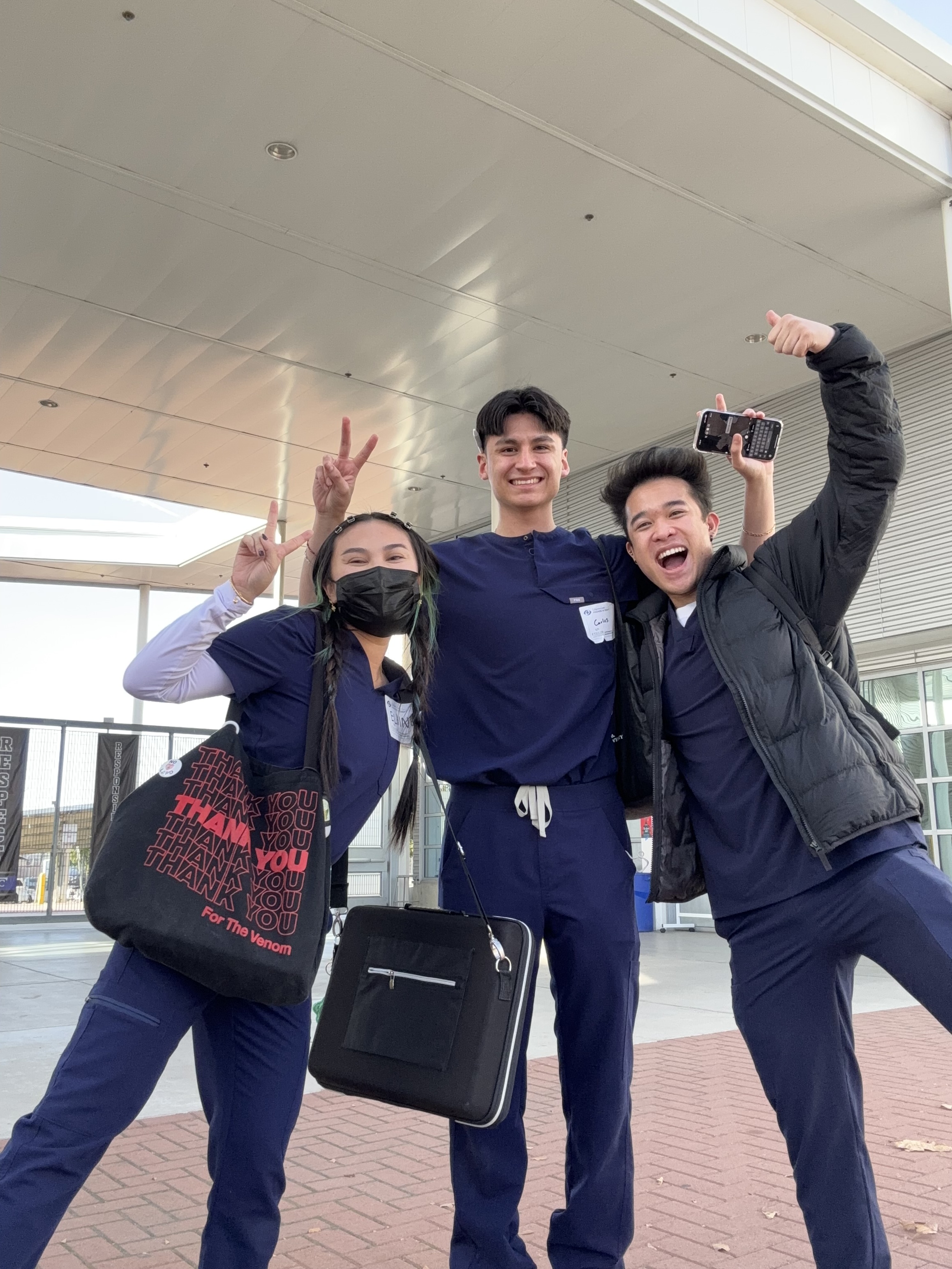 Three students wearing scrubs, smiling and making peace signs. 