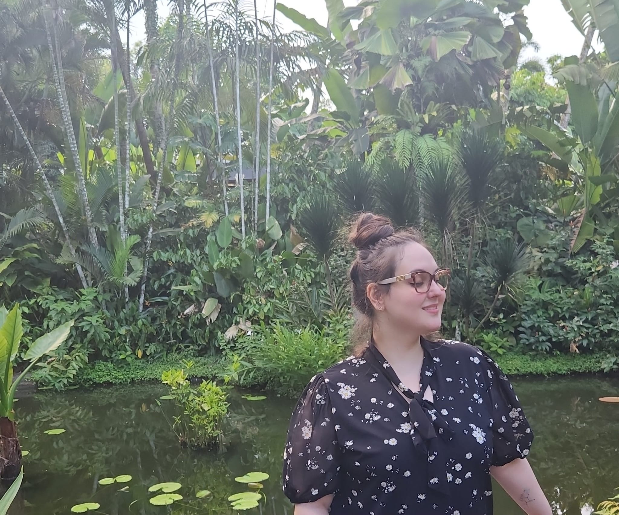 Picture of a person smiling with eyes closed in front of a background of green plants, trees, and a body of water.